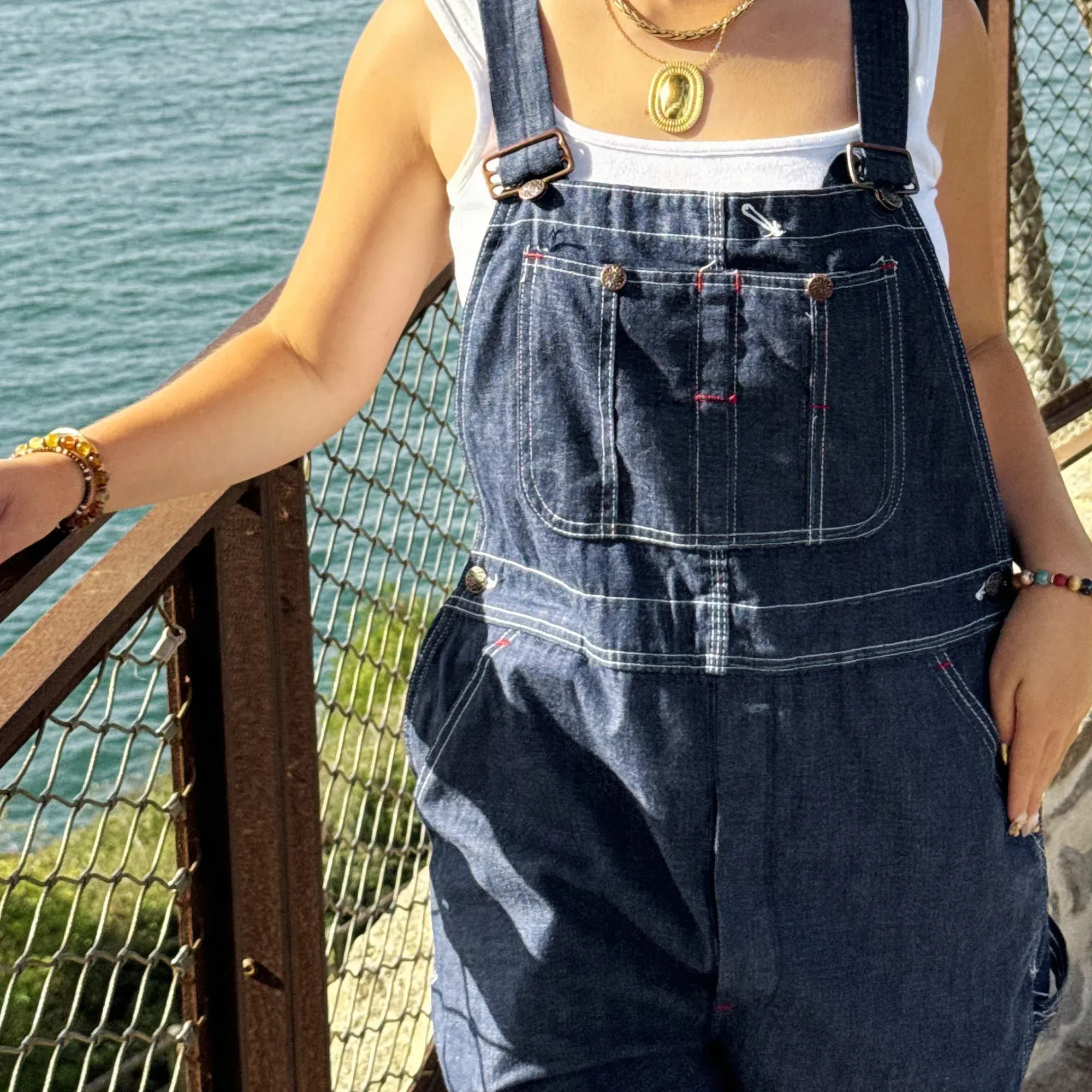 Salopette en denim portée devant un grillage, au bord de la mer